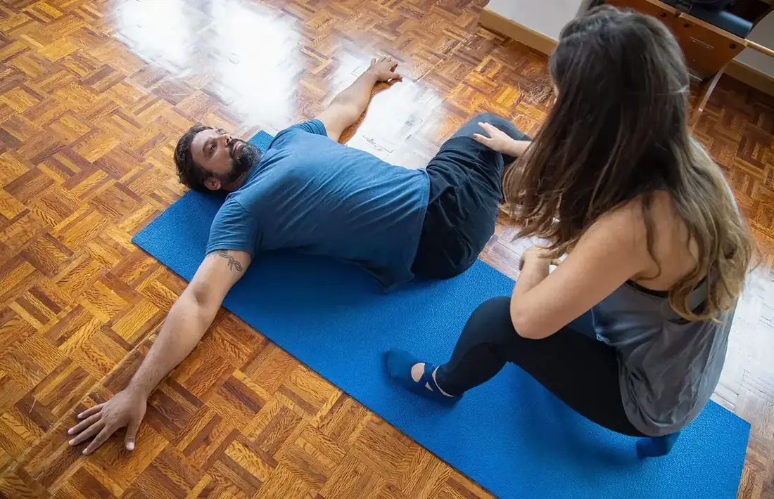 One person lying on a blue mat on a wooden floor while another kneels beside them